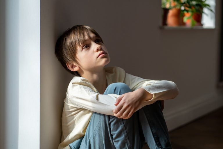 Lonely boy sadly looking up hugging knees sits near window at home alone. Kid psychological trauma